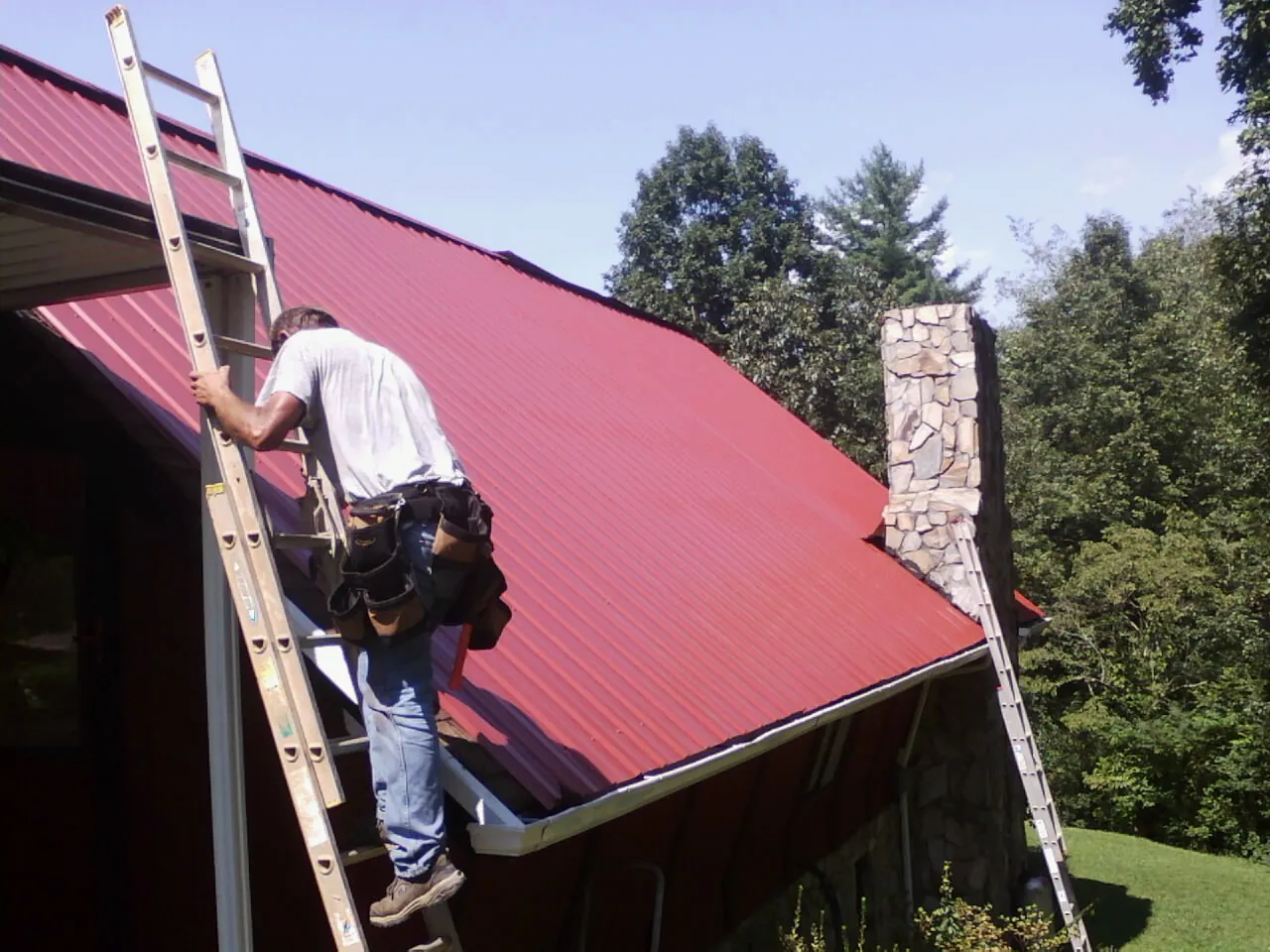 Roofer on a ladder beside a red metal roof and stone chimney — Soffit & Fascia Repair by Roofing Brothers in Nehalem